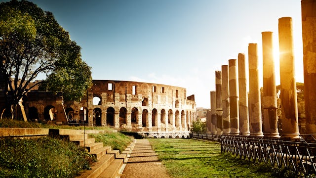 Het Colosseum bij zonsondergang met een pad omringd door oude zuilen en groene bomen.