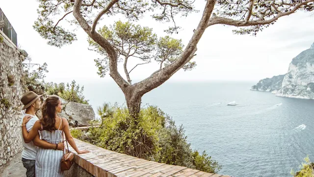 Twee reizigers wandelen langs een kustpad met uitzicht op zee en rotsachtige kliffen, omringd door mediterrane natuur en een helder uitzicht over het water.