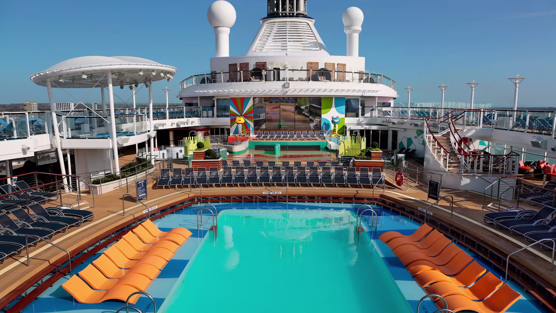 Zwembad op het dek van een cruiseschip met ligbedden, zonnedek en een groot scherm voor entertainment aan boord