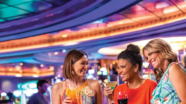 Drie gasten genieten van cocktails in een levendige bar aan boord van een cruiseschip met kleurrijke verlichting en een gezellige sfeer.