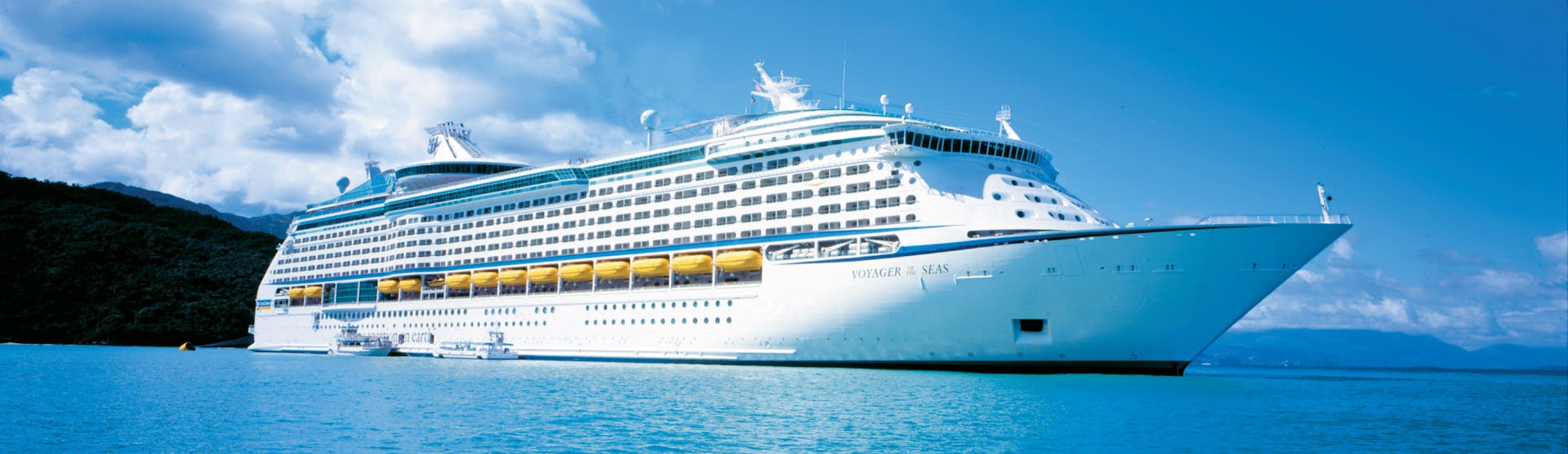 Luxe cruiseship op helderblauw water met een tropisch landschap en een deels bewolkte heel op de achtergrond.