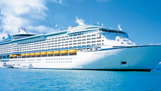 Luxe cruiseship op helderblauw water met een tropisch landschap en een deels bewolkte heel op de achtergrond.