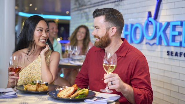 IC, Icon of the Seas, couple enjoying dinner at Hooked Seafood, smiling, wineglasses, enjoyment, Hooked Seafood signage on wall behind,