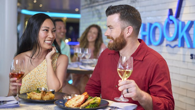 IC, Icon of the Seas, couple enjoying dinner at Hooked Seafood, smiling, wineglasses, enjoyment, Hooked Seafood signage on wall behind,
