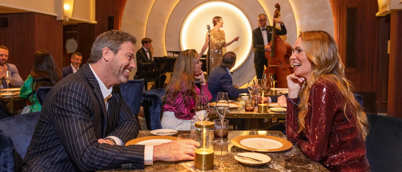 IC, Couple dining at Empire Supper Club onboard Icon of the Seas, musical group on stage in the background, nightlife, fun, evening, 1930s old New York theme, glamour,