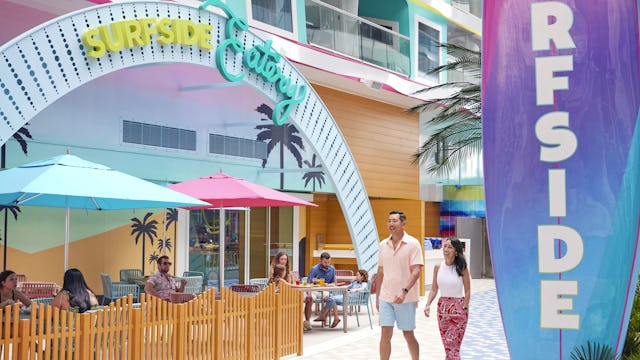 IC, Icon of the Seas, couple walking by entrance to Surfside Eatery, signage, exterior, guests sitting at tables, balconies above,