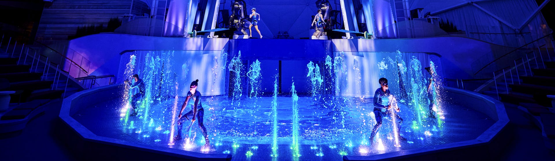 IC, Icon of the Seas, wide shot of performers, on stage in AquaTheater show. Aqua Action, fountains of water bubbling up, excitement, exciting, blue colors, stage lights,