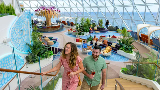 Stel geniet van drankjes in een lichte lounge op een cruiseschip met tropische beplanting, zitplekken en weids uitzicht over zee.