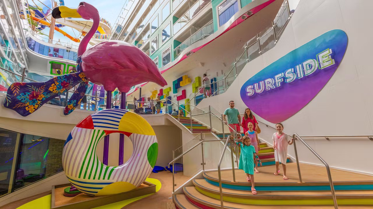 Kinderen en ouders lopen over een kleurrijk dek op een cruiseschip langs een grote flamingo en een Surfside-speelruimte. 