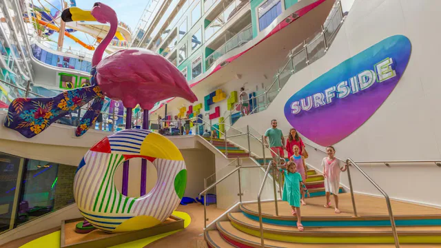 Kinderen en ouders lopen over een kleurrijk dek op een cruiseschip langs een grote flamingo en een Surfside-speelruimte.