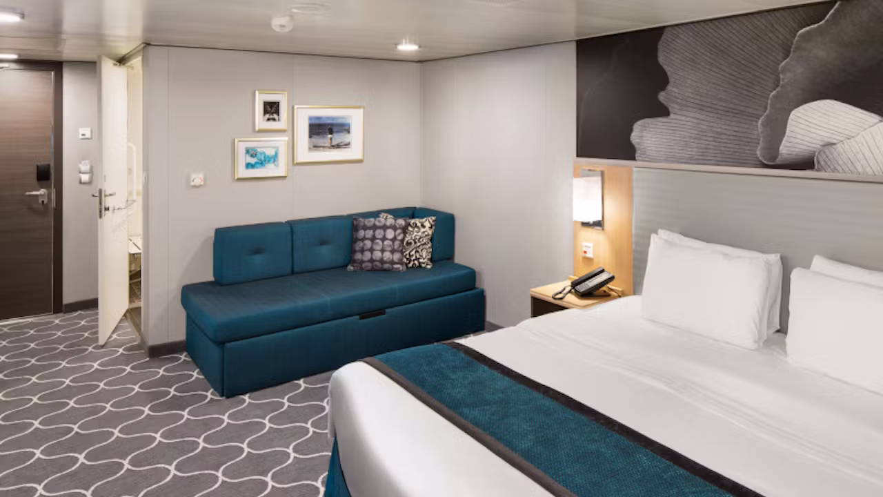 Moderne cruisehut met een tweepersoonsbed, blauwe bank, nachtkastje en zachte verlichting aan boord van een cruiseschip. 