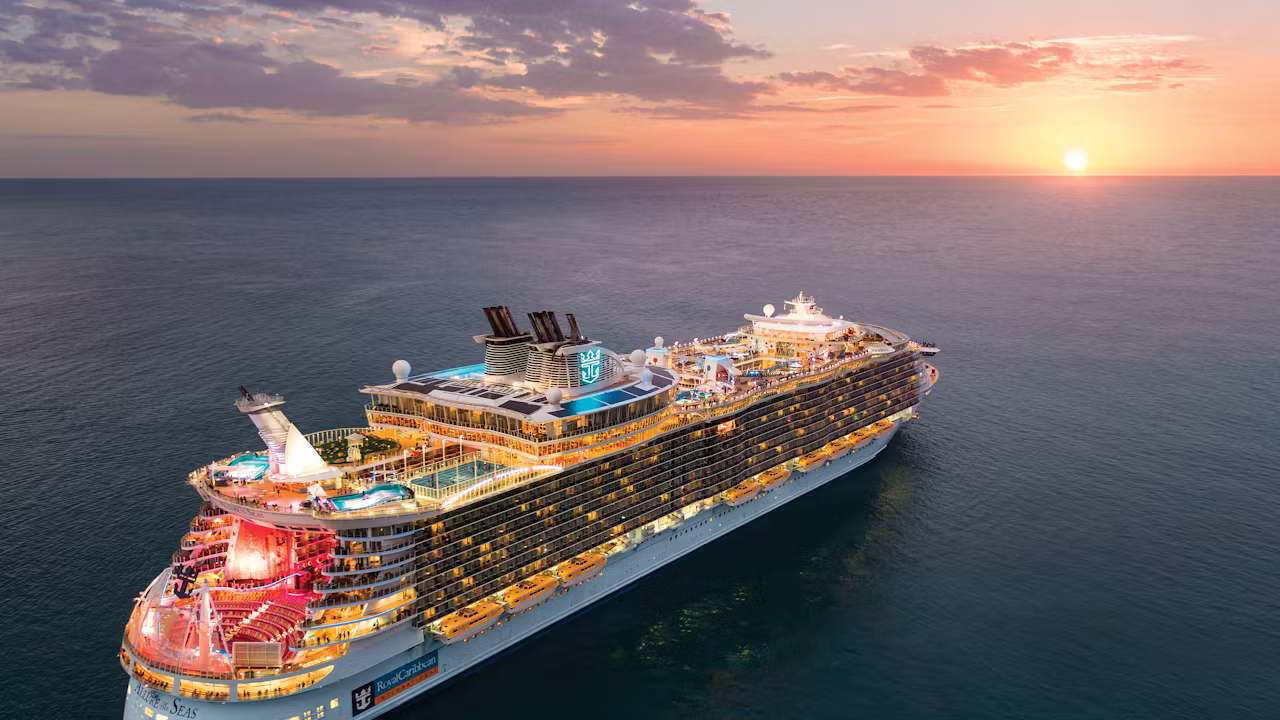 Verlicht Royal Caribbean-cruiseschip vaart bij zonsondergang over een kalme zee, met een kleurrijke lucht en uitzicht op het zonnedek. 