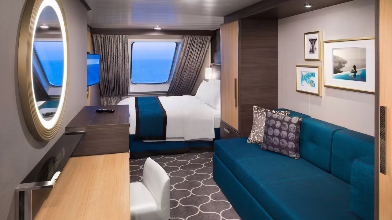 Moderne cruisehut met een tweepersoonsbed, blauwe bank, bureau, televisie en groot raam met uitzicht op zee. 