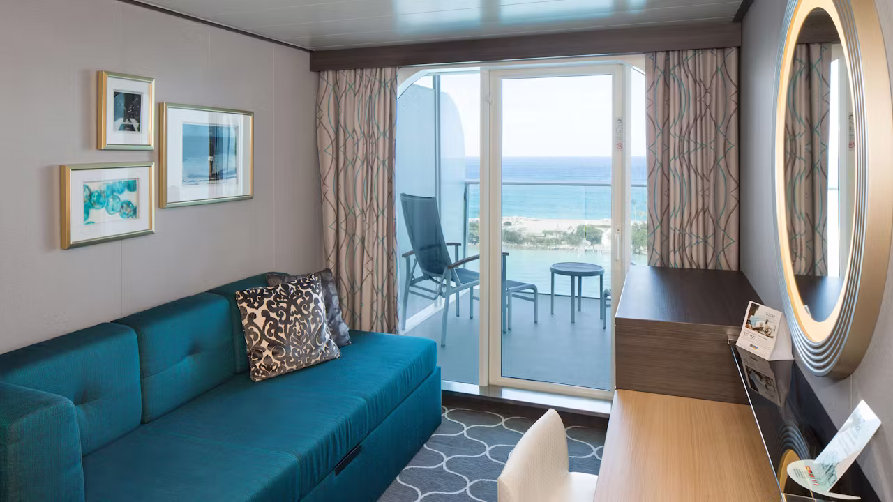 Balkonhut op een cruiseschip met blauwe bank, bureau, ronde spiegel en privébalkon met stoelen en uitzicht op zee. 