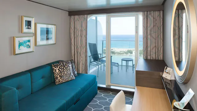 Balkonhut op een cruiseschip met blauwe bank, bureau, ronde spiegel en privébalkon met stoelen en uitzicht op zee.