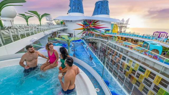 Vier gasten ontspannen met drankjes in een bubbelbad op het zonnedek van een cruiseschip, met uitzicht op het zwembad en kleurrijke waterattracties.