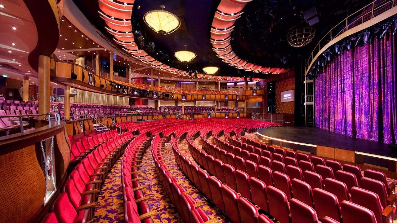 Groot theater op een cruiseschip met rode stoelen, een ruim podium en sfeervolle verlichting voor live shows en entertainment. 