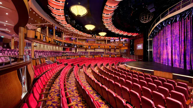 Groot theater op een cruiseschip met rode stoelen, een ruim podium en sfeervolle verlichting voor live shows en entertainment.